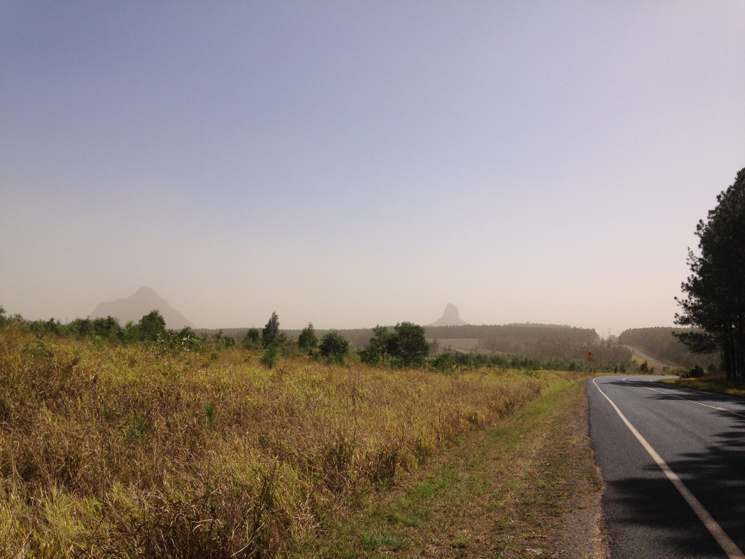 Vista from the road. Some road surface on the right, on the left is open field of brown grass. Distance has mountains (identifiable as Glashouse Mountains). The atmoshpehere is obviously hazy and slightly red brown with dust.