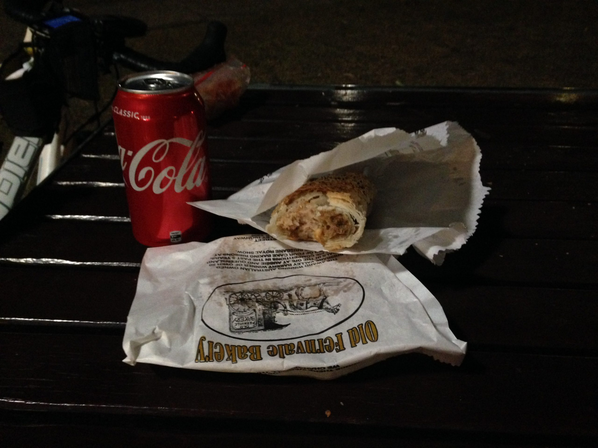 A dark table.  A red can of 'Coke', an open paper bag with a sausage roll, and a closed paper bag with the Fernvale Bakery artwork.